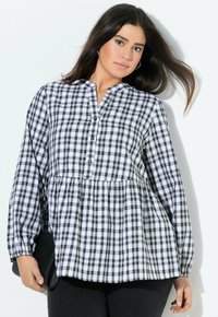 Frau mit langen dunklen Haaren, die eine schwarz-weiße karierte Bluse trägt und eine schwarze Clutch vor einem weißen Hintergrund hält.
