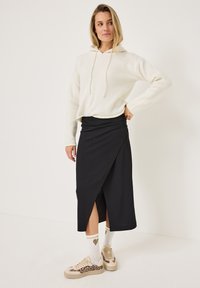 Zwarte wikkelrok met een zijsplit, gecombineerd met een witte hoodie. Kenmerkt zich door vlakke naden, beige sneakers en gestreepte sokken met harten.