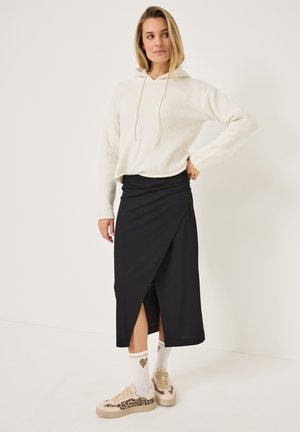 Jupe noire portefeuille avec une fente sur le côté, associée à un sweat à capuche blanc. Présente des coutures à plat, des baskets beiges et des chaussettes à motifs avec des cœurs.