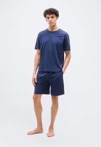 Νέος άντρας όρθιος, ξυπόλητος, φοράει μπλε κοντομάνικη μπλούζα και ταιριαστό σορτς, μπροστά σε ουδέτερο φόντο.