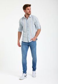 Camicia azzurra chiara con righe verticali, abbinata a jeans blu aderenti e sneakers bianche. La camicia presenta un colletto e maniche arrotolate.