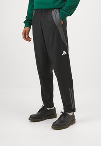 Pantalones deportivos negros con un ajuste ajustado, paneles laterales grises elegantes y logo de Adidas en blanco; combinados con zapatos negros y calcetines grises.