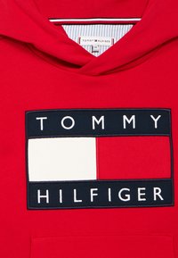 Κόκκινο βαμβακερό φούτερ με κουκούλα που διαθέτει μεγάλο κεντημένο λογότυπο Tommy Hilfiger. Το λογότυπο περιλαμβάνει ένα ναυτικό ορθογώνιο κολάρο με λευκές και κόκκινες λεπτομέρειες.