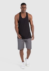 Camiseta sin mangas negra con escote redondeado, combinada con pantalones cortos grises. El modelo lleva zapatillas deportivas blancas. Fondo liso.