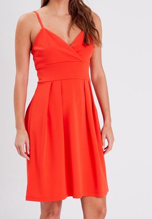 Femme portant une robe rouge sans manches, d'une longueur au genou, avec un décolleté en V et une taille cintrée, debout devant un fond gris clair uni.