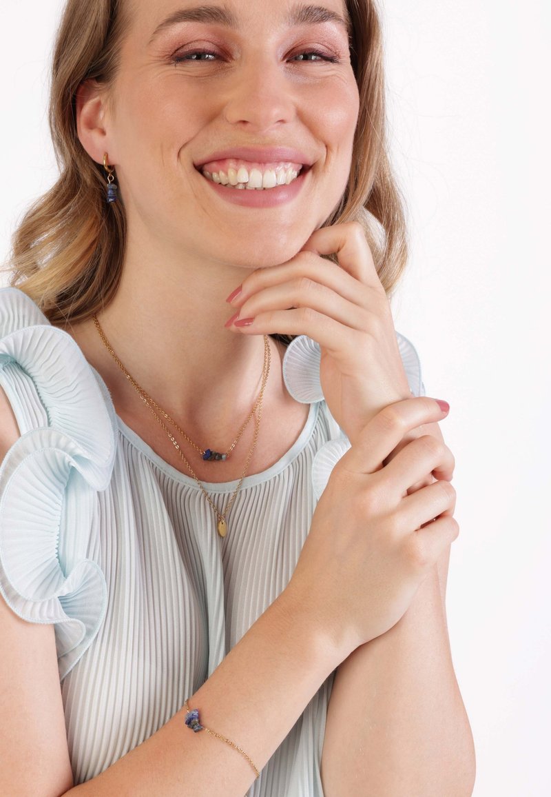 Gouden sieraden met een gelaagde ketting met blauwe edelstenen en een ovale hanger, vergezeld van bijpassende oorbellen. Het model draagt een lichtblauw geplooid topje.