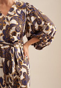 Fließendes Kleid in Weiß mit blauen und goldenen Blumenmustern. Es hat einen tiefen V-Ausschnitt und voluminöse Puffärmel, die an der Taille mit einem Gürtel gerafft sind.