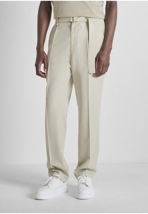 Uomo che indossa pantaloni beige a vita alta con cintura in tessuto abbinata e sneaker bianche in pelle, in piedi contro uno sfondo semplice.