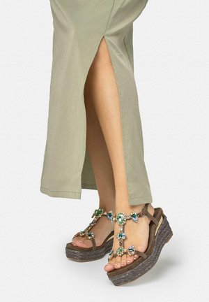 Piernas con una falda midi verde claro con abertura lateral, llevando sandalias de plataforma marrones adornadas con grandes gemas verdes y azules.