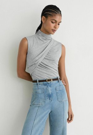 Vrouw met gevlochten haar, die een mouwloze grijze coltop draagt en lichtblauwe jeans met hoge taille, voorzakken en een bruine riem.