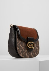 Borsa a tracolla in suede e pelle marrone con una chiusura ovale dorata e un motivo di carrozza a cavallo sulla parte anteriore. Forma arrotondata con accenti neri.