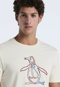 Crèmekleurig katoenen t-shirt met een pinguïnontwerp omlijnd in rode, blauwe en beige borduursels. Het shirt heeft een klassieke ronde hals.