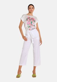 Weißes, blumiges Grafik-T-Shirt mit kurzen Ärmeln, kombiniert mit hochgeschnittenen weißen Leinenhosen. Das Modell trägt grüne Riemchenschuhe.