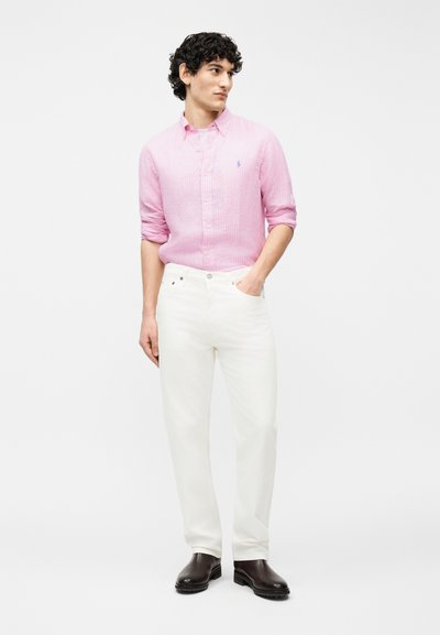 Άνδρας που φοράει ροζ πουκάμισο με κουμπιά, άσπρο παντελόνι, καφέ μπότες, στέκεται με το ένα χέρι στην τσέπη, κοιτώντας προς τα πλάγια σε μονόχρωμο φόντο.