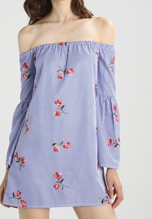 Femme portant une robe à rayures bleues et blanches, épaules dénudées, manches longues et motifs floraux rouges partout.