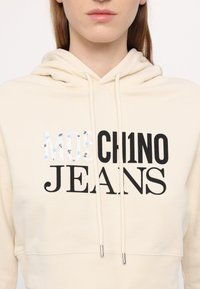 Krémová mikina s kapucňou a šnúrkou, na ktorej je nápis "MOSCHINO JEANS" v rôznych písmach. Text obsahuje kvetinové akcenty v modrej a čiernej farbe.