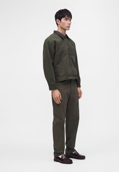 Jeune homme debout, portant une veste et un pantalon vert foncé avec des chaussures en cuir brun foncé sur un fond clair uni.