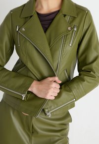 Personne portant une veste en cuir vert olive avec des zips en argent, en train de fermer le zip avant, associée à un pantalon assorti.