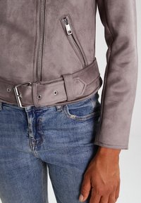 Giacca in suede grigia con chiusura a zip, dettagli con cintura a scatto e superficie liscia, abbinata a jeans in denim blu. Vista ravvicinata della vita.