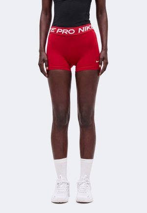 Femme portant un short Nike Pro rouge, un haut noir, des chaussettes blanches et des baskets Nike blanches, debout devant un fond uni.