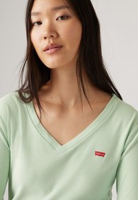 Duga majica s dugim rukavima u mint zelenoj boji s V-izrezom. Ima mali crveni Levi's logo s lijeve strane grudi. Mekan materijal.