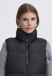 Zwarte puffer-vest met hoge kraag, rits aan de voorkant en grijze mouwen van een trui. Gladde textuur, gewatteerd patroon en zilverkleurige metalen details.