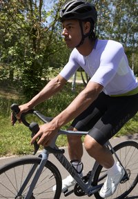Maillot de cyclisme violet clair à manches courtes, cuissards de cyclisme noirs avec motif hexagonal, et chaussures de cyclisme blanches. En train de faire du vélo sur un vélo gris.