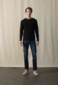 Sort langærmet skjorte parret med blå jeans og beige sneakers. Skjorten har et lille logo på brystet. Enkel, afslappet påklædning.