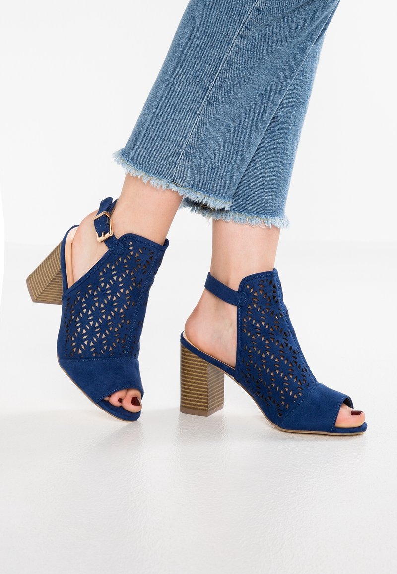 Sandales à talons ouvertes en daim perforé bleu, avec un talon bloc empilé et une bride à la cheville. Le design comprend des découpes florales et une finition lisse.