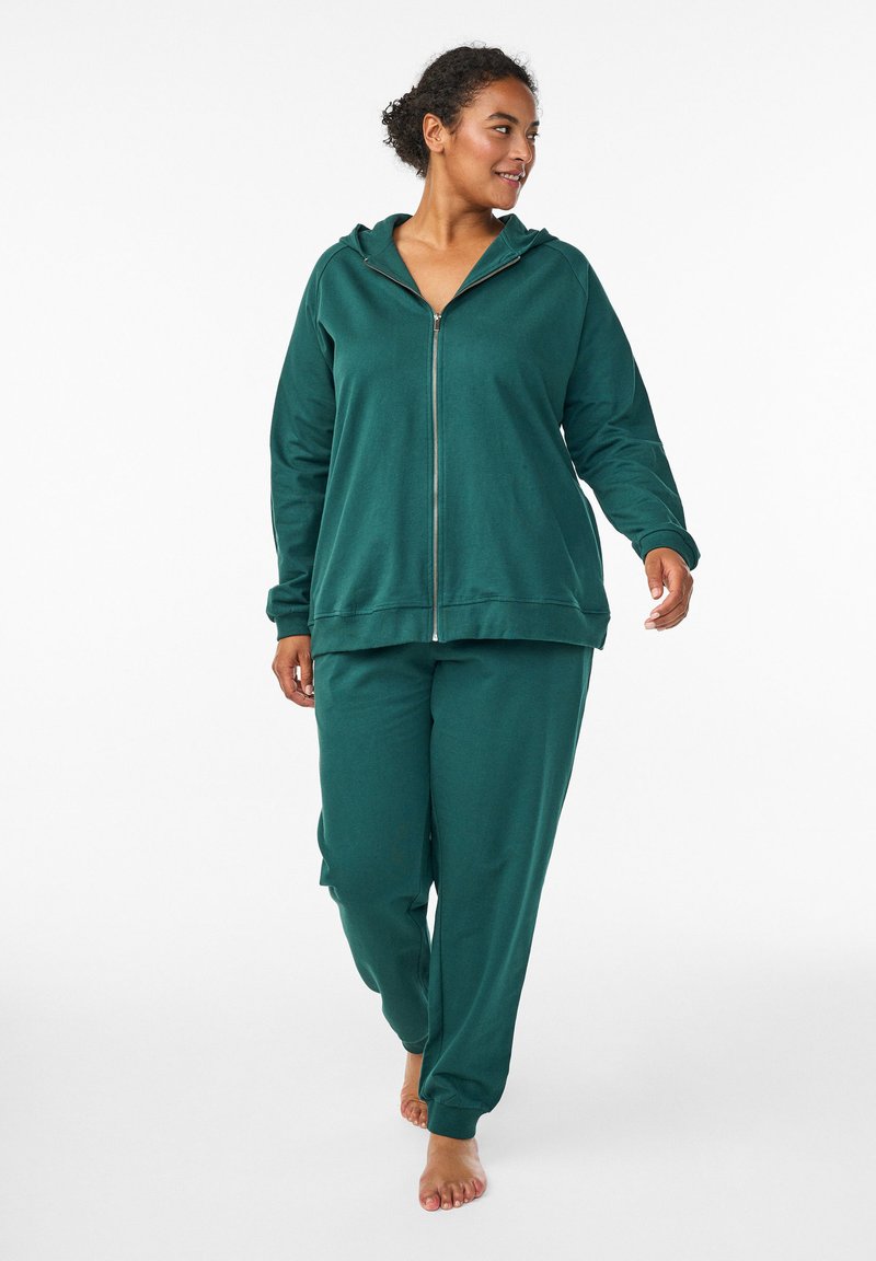 Grüne Kapuzenjacke mit Reißverschluss aus weichem Material, kombiniert mit passenden, locker sitzenden Hosen. Das Outfit hat entspannte Ärmel und Hosenabschlüsse.