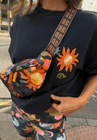 Musta sweatshirt, jossa on kirjailtu auringon kuvio, värikäs kuvioitu vyölaukku ja eloisa kukkakuvioiset leggingsit. Vyölaukussa säädettävä hihna.