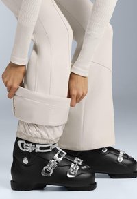 Czarne buty narciarskie z srebrnymi klamrami, w połączeniu z beżowymi spodniami narciarskimi z marszczonym mankietem. Teksturowana tkanina spodni dodaje detali.
