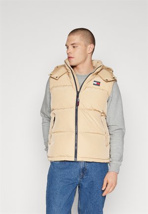 Beige puffer vest with a zipped front, collar, and side pockets. Features a logo patch, quilted design, and is worn over a gray long-sleeve shirt.