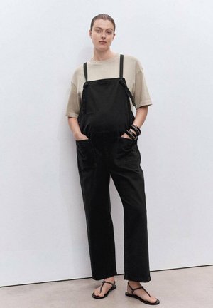 Zwarte denim overall met verstelbare schouderbanden, zijzakken, losse pasvorm, gecombineerd met een beige T-shirt met korte mouwen en platte sandalen.