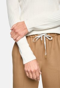 Weißes, geripptes Langarmoberteil mit engen Bündchen, kombiniert mit hellbraunen Hosen mit Kordelzug und goldenen Akzenten.