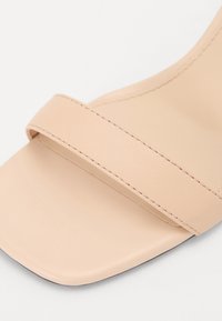 Sandale beige à bout ouvert avec une large bride unique et une partie avant de forme carrée, montrant des coutures détaillées sur un matériau souple.