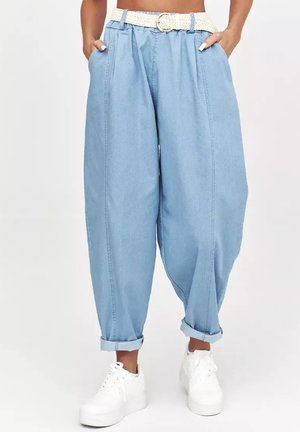Jasnoniebieskie jeansy damskie z wysokim stanem, luźnym krojem, białym plecionym paskiem, podwiniętymi nogawkami, zestawione z białymi sneakersami na platformie. Ręce w kieszeniach.