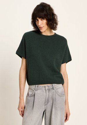 Frau mit kurzem, lockigem, dunklem Haar, die einen dunkelgrünen, kurzärmligen Pullover und hellgraue, gefaltete Jeans trägt, vor einem hellen Hintergrund.