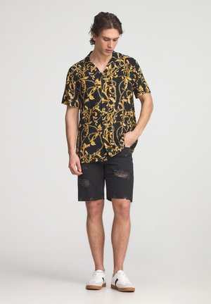 Maglietta nera a maniche corte con stampa floreale gialla e catena, abbinata a pantaloncini neri strappati e sneakers bianche con suola beige.