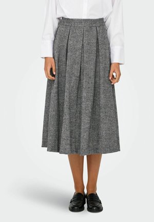 Femme portant une jupe midi plissée grise, une chemise blanche à manches longues et des mocassins noirs, debout devant un fond clair uni.