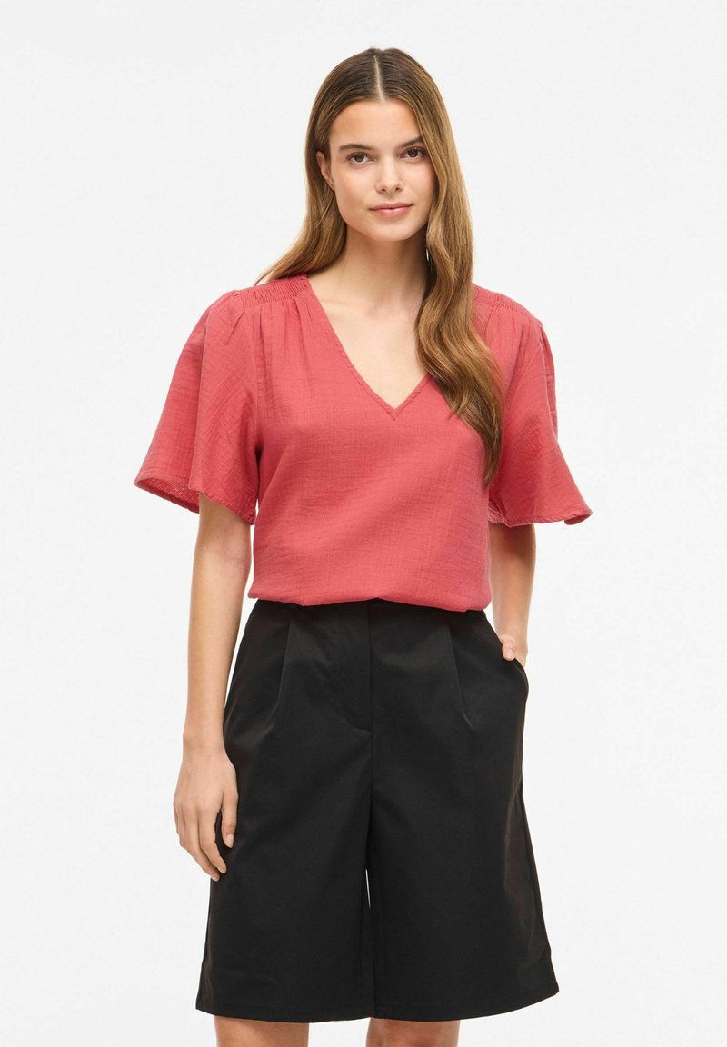 Femme aux longs cheveux bruns portant un chemisier corail à col en V avec des manches volantes et un short noir taille haute à jambes larges, debout avec une main dans la poche.