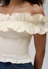 Blusa con hombros al aire en suave tejido crema con escote con volantes y fruncido texturizado. Terminada con un dobladillo con volantes, ideal para combinar con vaqueros.