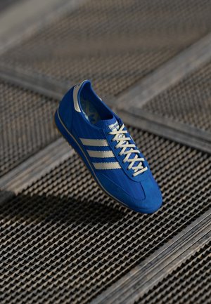Blå Adidas sneaker med hvide striber og snørebånd, svævende over en tekstureret metalristgulv.