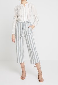 Witte blouse met verticale kantdetails, gecombineerd met blauw-witte gestreepte, hoog-getailleerde cropped broek en beige sandalen met hak en banden.