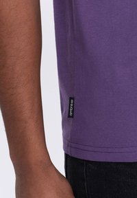 Maglietta viola in cotone, con etichetta laterale con il marchio "ambre", orlo cucito e texture liscia. Dettaglio del braccio visibile nell'inquadratura.