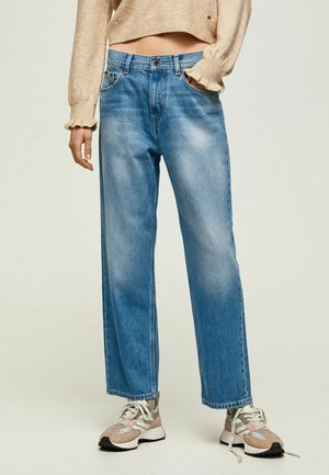 Kobieta ubrana w jasnoniebieskie jeansy o wysokim stanie i prostych nogawkach, beżowy krótki sweter oraz beżowo-różowo-szare trampki, stojąca z jedną ręką w kieszeni.