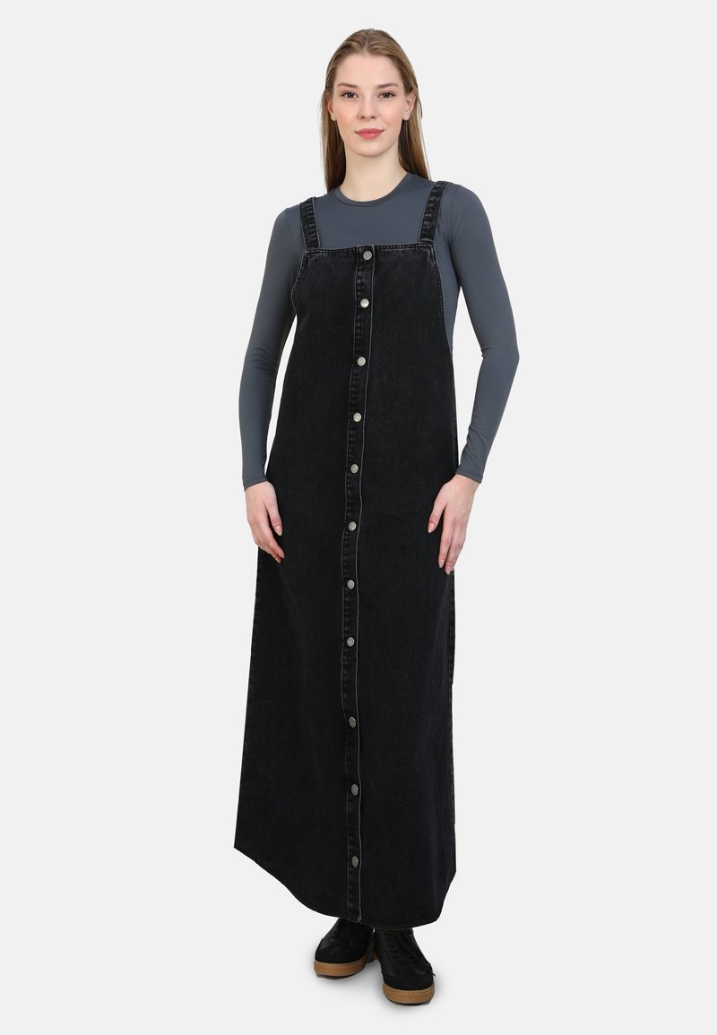 Salopette in denim nero con taglio dritto, chiusura frontale con bottoni, spacchi laterali e bretelle regolabili. Abbinata a una maglietta a maniche lunghe grigia.