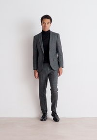 Completo grigio a fantasia con vestibilità sartoriale, abbinato a un dolcevita nero. Presenta eleganti scarpe nere e uno sfondo minimalista.