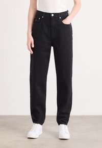 Personne portant un jean noir taille haute coupe décontractée, baskets blanches et haut blanc, debout sur un sol uni devant un mur blanc.