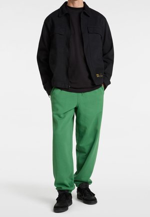 Zwarte jas met twee borstzakken, gedragen over een zwart shirt. Groene joggingbroek met een losse pasvorm, gecombineerd met zwarte sneakers.